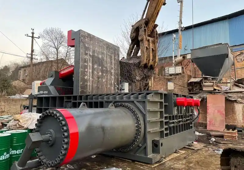 Material-handler grab collecting a compacted steel-scrap bale beside a baler
